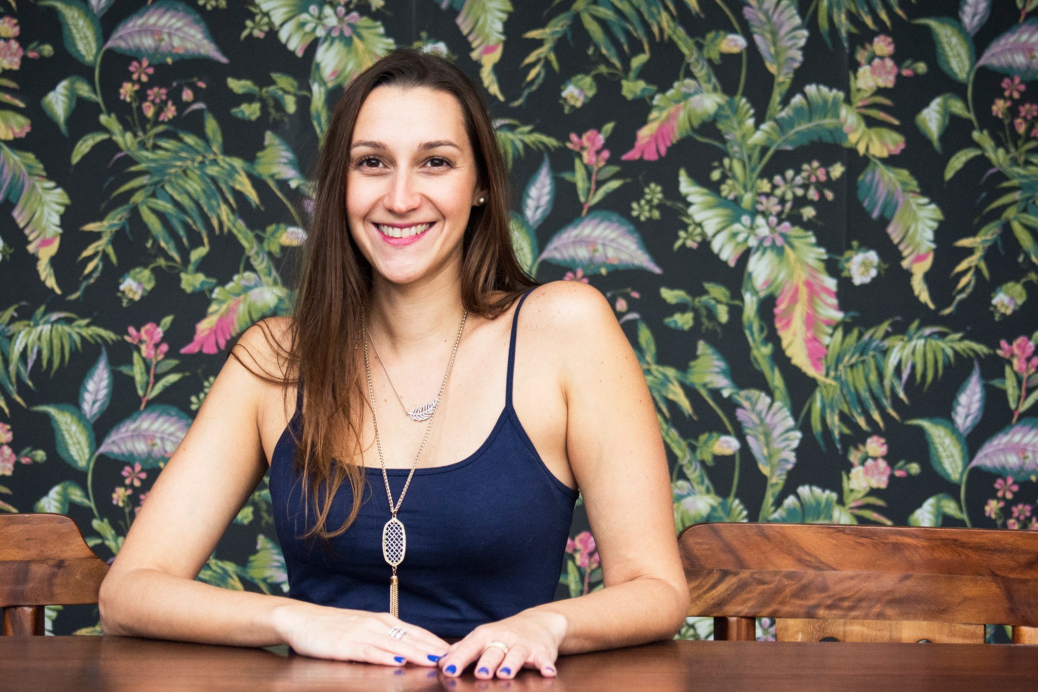 Giannina Cappella smiling at a wooden table in front of tropical botanical wallpaper